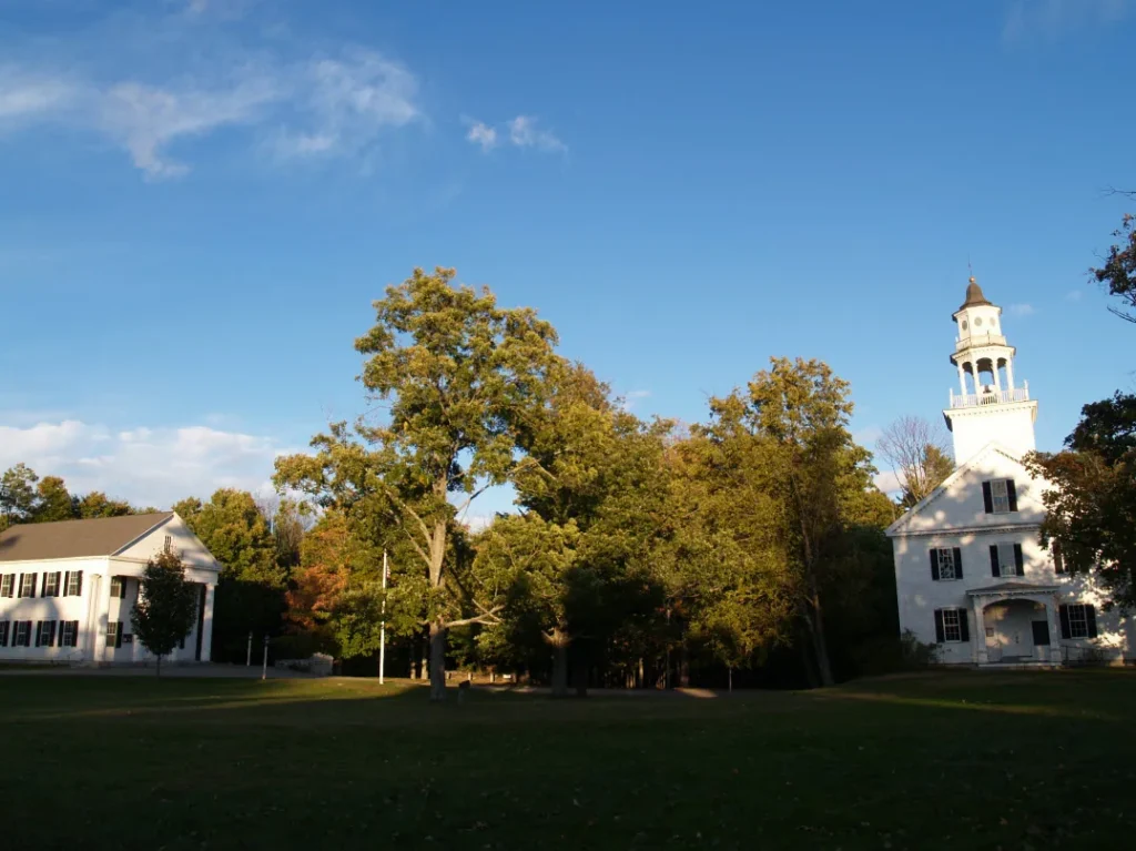 Campus Scene White Two Story Building on the Left a Tall White Church Steeple on the Right with Trees and a Grassy Lawn